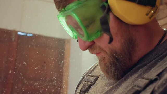 Close up shows part of man face wearing green safety glasses and yellow ear protection with wood dust particles visible in air. scene is indoors likely during woodworking or carpentry