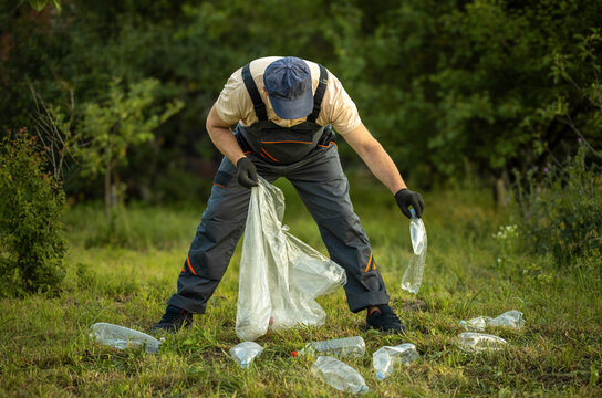 Worker collects discarded plastic bottles in garden, holding one above large clear waste bag. Gloved hands gather litter from grass, supporting outdoor cleanup and environmental care.