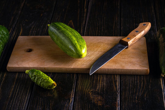 Fresh cucumber and kitchen knife rest on wooden board. Whole green vegetable and small pickling cucumber are set on rustic dark tabletop, suggesting simple home cooking preparation.