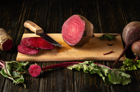 Fresh beetroot sits sliced on wooden cutting board beside uncut roots. Kitchen knife and scattered leaves emphasize rustic food preparation, vivid texture, and deep crimson color beside round roots.
