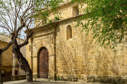 Church of Santo Domingo in Ubeda, Spain, showcasing its intricately carved plateresque doorway and robust stone facade under sunlight, with a prominent tree framing the historic architecture