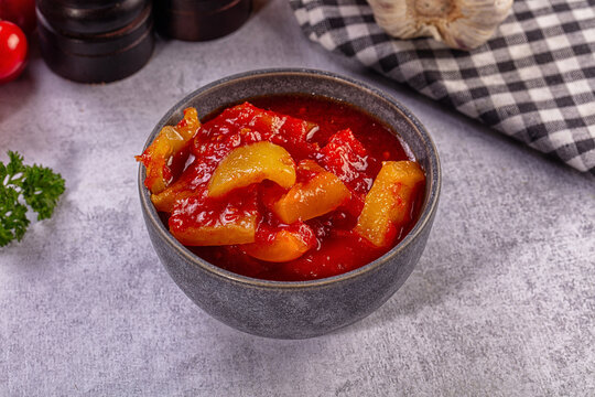 Bowl of lecho with red tomato sauce and yellow bell peppers on grey surface beside checkered cloth and garlic