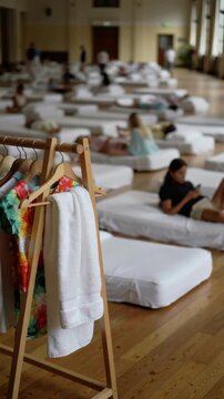 Wooden clothes rack with white towel and floral dress in a large room with people on floor mattresses