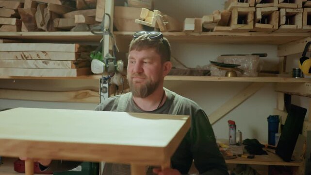 Man inspects large unfinished wooden table top in his workshop surrounded by woodworking tools like saws. Man lifts tabletop to examine joinery details while shelves filled with lumber are