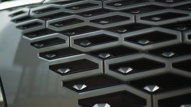 Close up view of vehicle front end focusing on geometric grille with hexagonal patterned metalwork headlight assembly visible. Camera slowly pans across intricate texture details reflections