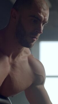 Focused muscular athlete training shirtless on exercise equipment in a dimly lit gym, highlighting powerful shoulders, arms, and upper body definition during intense workout