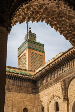 Low-angle perspective framing the ornate minaret and courtyard architecture of the historic Al-Attarine Madrasa through an intricately carved Moorish archway in Fes, Morocco