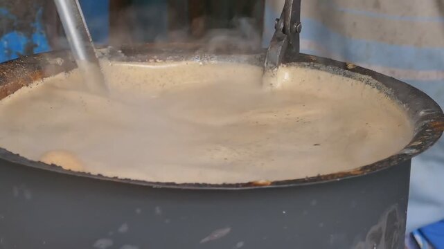 Creamy thick batter swirls inside large metal mixer pot as steam rises from rusted rim in blue industrial workshop, heavy production equipment close up
