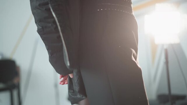Close up shows bottom half of standing woman focusing on her legs and hands as woman adjusts clothing in front of bright studio lights. background includes professional photography equipment