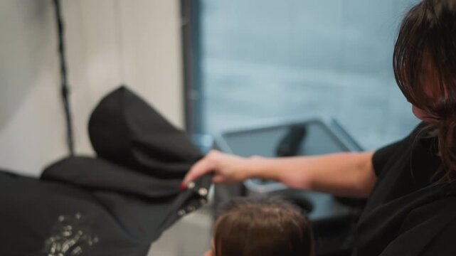 Close up view of woman as woman adjusts black hair cape on another woman seated at salon chair. stylist hands are visible carefully fastening buttons to secure client clothing before