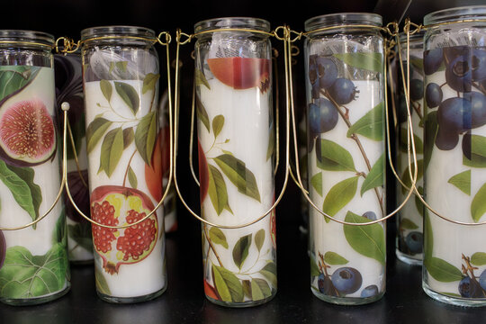 Decorative glass candles printed with colorful fruit designs like fig, pomegranate and grapes, neatly arranged together on a shelf with gold decor, film grain, high quality photo