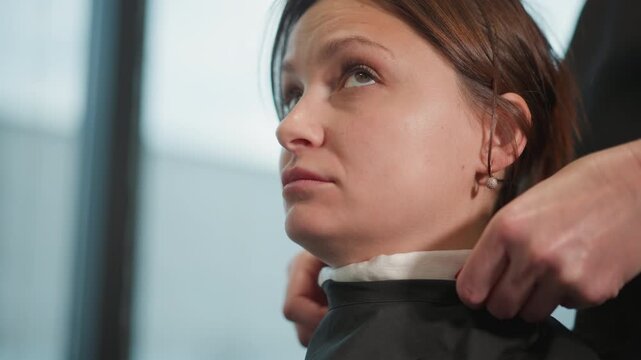 Woman sits calmly as skilled professional hands adjust her dark brown hair into black salon cape. close up emphasizes careful interaction between stylist capable red painted nails and client