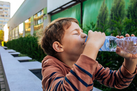 Young boy in a striped hoodie drinking cold water from a plastic bottle on a hot sunny summer day outdoors, child thirst and hydration awareness concept, film grain, high quality photo
