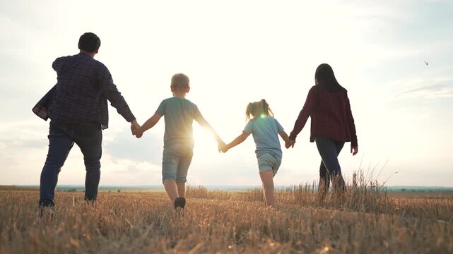 Family holding hands running together in field at sunset. Parents and children run while holding hands outdoors. Family runs across countryside field holding hands at golden sunset time.