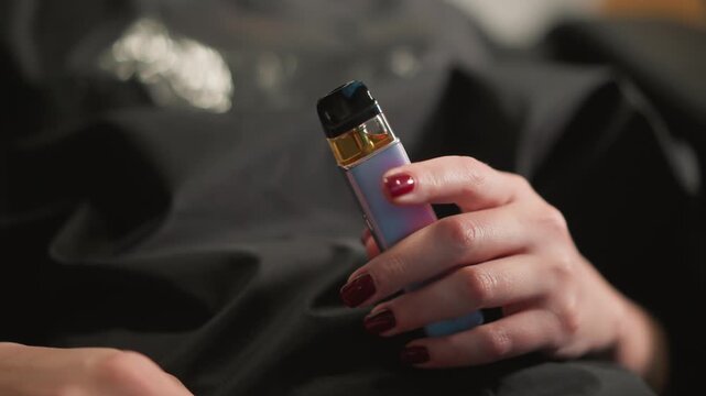 Close up shows woman manicured nails gripping small electronic vaporizer. focus remains steady highlighting details of hand and device against dark background