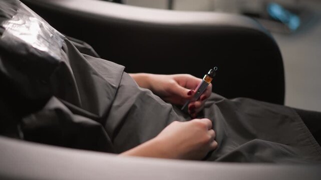 Client with red nail polish holds electronic vaping device between fingers and rests on lap. Salon worker gloved hands perform hair treatment services like cutting or coloring in background