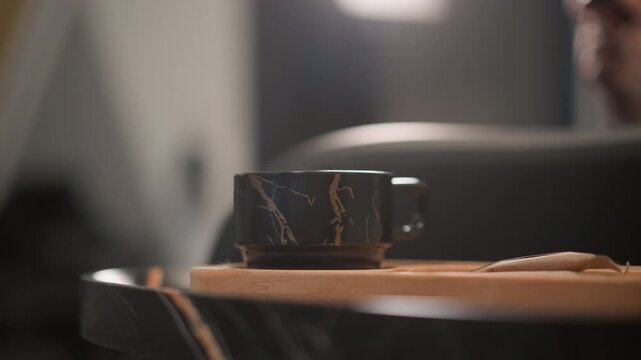 Steady shot focuses on dark marble patterned ceramic cup placed centrally on light wood serving board. background remains softly blurred with warm ambient lights emphasizing texture