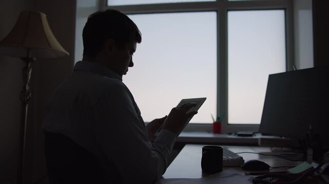 Silhouette of person touching tablet screen at backlit office desk beside glowing window with pale sky gradient in quiet remote workspace low light