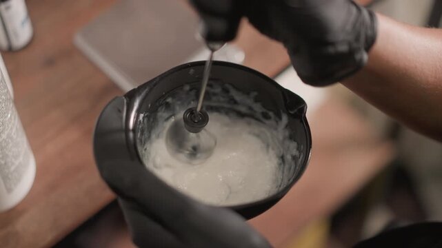 Close up view of hand wearing black glove pouring liquid into mixing bowl with electric mixer in motion indicating hair color preparation. scene shows careful addition of product to ensure