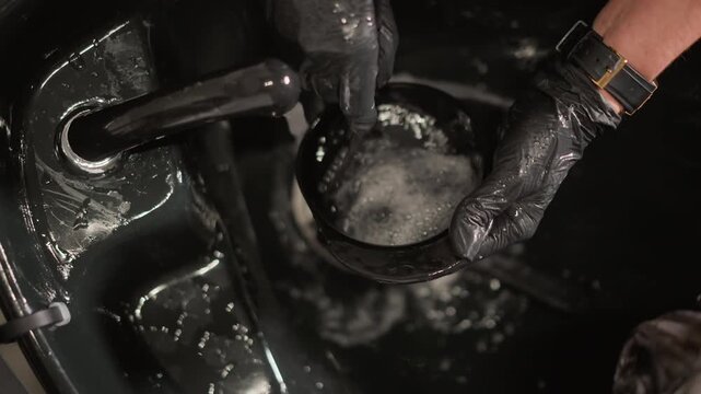 Close up on gloved hand holding and rinsing object in soapy sink. Gloved hands manipulate small container under running water likely part of hair or beauty treatment process