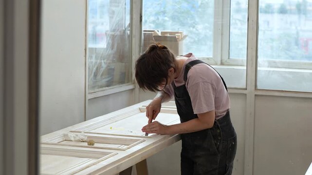 Concentrated skilled craftswoman restoring vintage wooden door using chisel tool in restoration studio, focused female crafter removing excesses of putty filler from antique door panel in workshop.