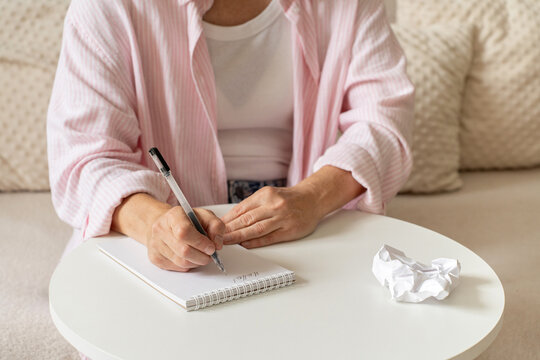 Mature woman writes in notebook at home, pen and crumpled sheet of paper lie on table, calm personal notes and measured daily life. A cozy, light-filled home setting