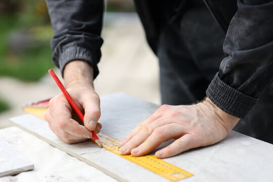 A person is using a red pencil to mark measurements on a piece of stone. They are holding a ruler with one hand and writing with the other. This is happening outdoors in a workshop area.