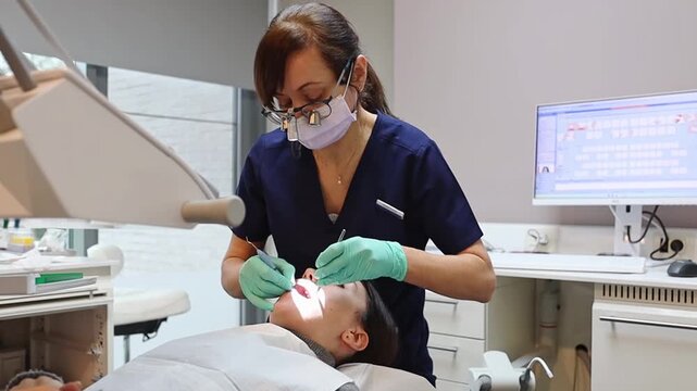 One young Caucasian female dentist in blue uniform and green gloves cleaning teeth from tartar and plaque with special instruments to girl patient, medium shot side view. Concept of gum and teeth trea