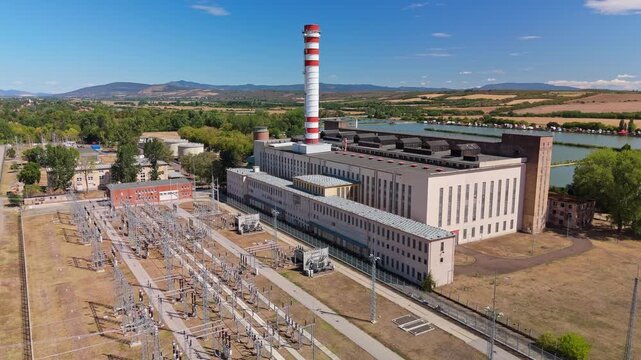 Drone view of a thermal power plant with a striped smokestack, electrical substation and cooling lake in Lőrinci, Hungary, surrounded by farmland and low hills.