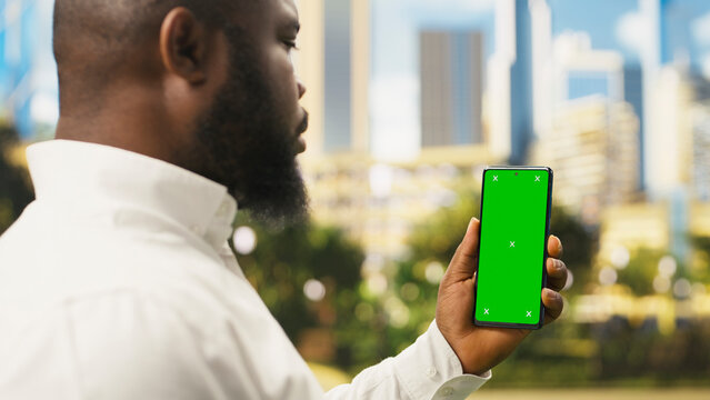 Vertical chroma key mockup on mobile phone held by man in urban park, providing replaceable display for website or advertising content.