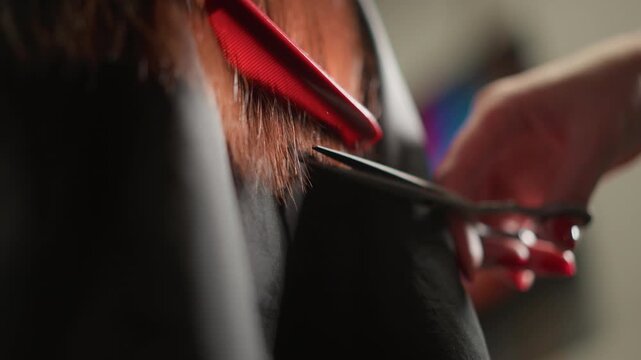 Close up shows person getting their reddish brown hair cut with visible strands falling as hand holds tools. red comb and scissors are used on client shoulder length locks while hands steady
