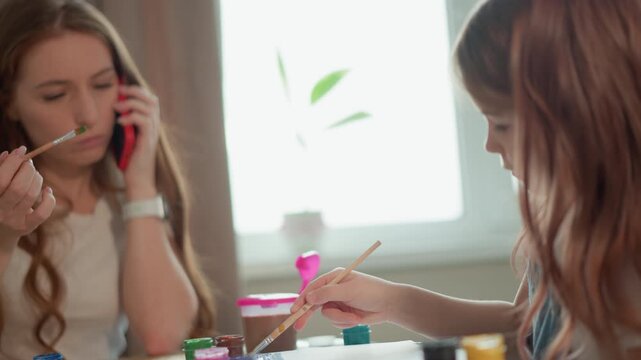 Mother talks on phone while sitting next to her daughter who is painting at table. They share space with paint containers visible suggesting family activity