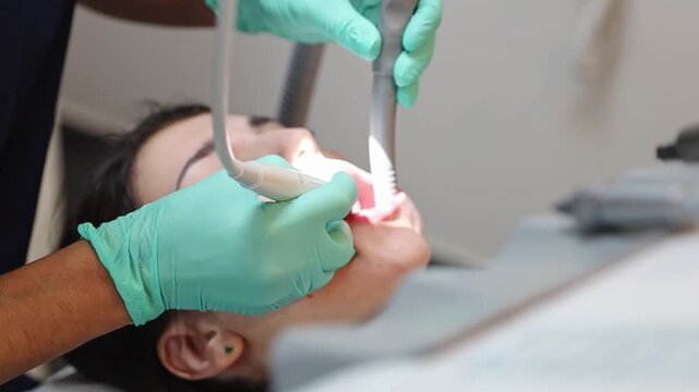 One young Caucasian unrecognizable female dentist in green gloves who cleans lower front teeth from tartar and plaque with special instruments to a girl patient, very close-up side view. Concept of gu