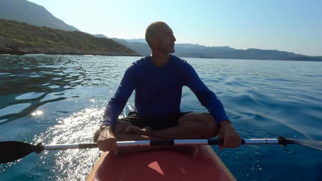 Tranquil man sitting in a meditative lotus pose on a red paddleboard, holding a paddle and peacefully floating on the calm blue sea with islands
