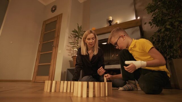 Loving mother and her son with a bandaged hand are playing together, patiently building a domino chain with wooden blocks on the floor in their cozy living room during a quiet evening