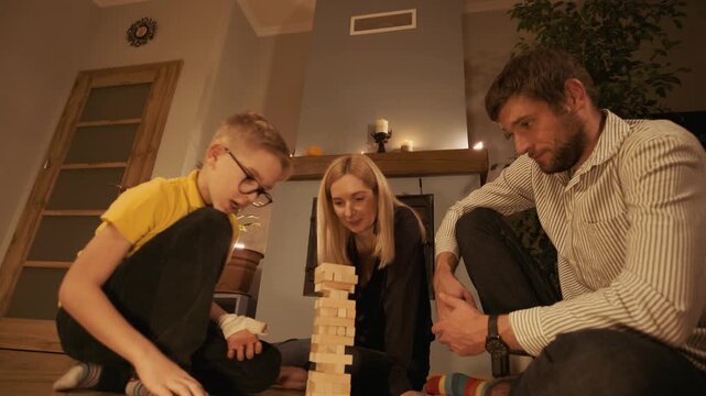 Happy caucasian family spending a cozy evening together playing a board game on the floor by the fireplace. Parents and their son building a tower from wooden blocks in a warm atmosphere