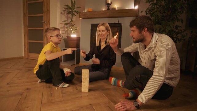 Adorable family with a young son spending a cozy evening together, sitting on the floor by a warm fireplace and playing rock paper scissors to decide who plays the wooden block tower game next