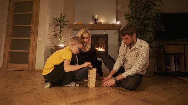 Lovely family with a child playing a wooden block tower game on the floor in their cozy living room, spending a warm and happy evening together near a burning fireplace at home