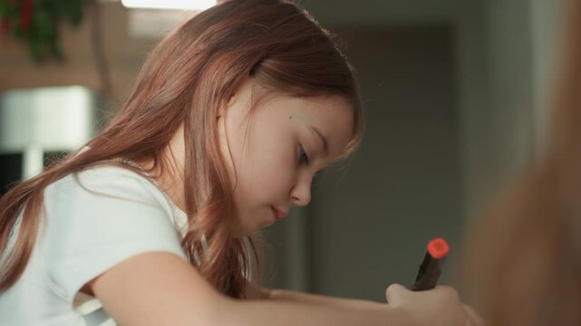 Young girl with long hair is deeply focused on drawing or coloring holding markers. scene takes place indoors under soft lighting emphasizing her concentration and creative activity