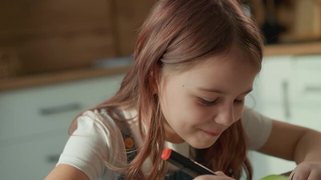 Young girl is deeply focused on cutting green paper using scissors indoors. Her hair detail and concentrated facial expressions are visible as girl works with domestic kitchen setting softly