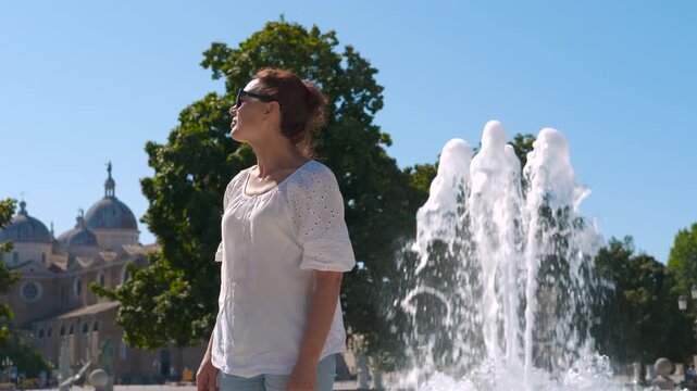 Tourist enjoying fountain in padua, italy, near the scrovegni chapel. Woman in white shirt and sunglasses relaxing near historic scrovegni chapel fountain, enjoying sunny summer day in padua, italy