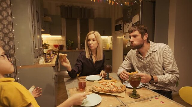 Joyful family with a young son celebrating and eating pizza together at the dinner table in their cozy kitchen, sharing a meal and enjoying quality time in a warm, welcoming atmosphere