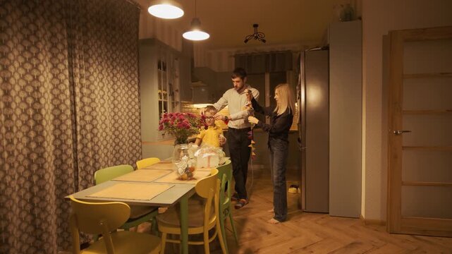 Joyful young parents with their little son preparing for a birthday party together, untangling a garland with colorful flags in a cozy kitchen decorated with flowers in the evening