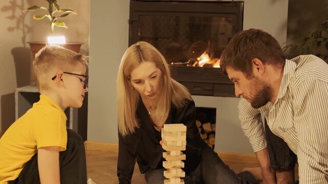 Lovely family with son playing a wood block stacking game together in the living room. Cozy evening with parents and child enjoying a board game near a warm fireplace at home