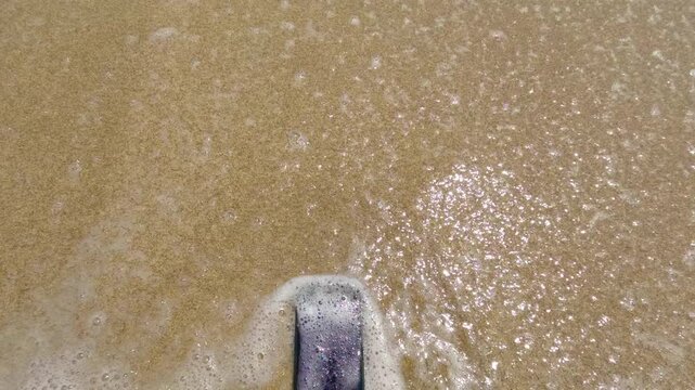 Forgotten modern smartphone lying on the golden sand of a beach as gentle sea waves wash over it, symbolizing loss, digital detox, or the fragility of technology