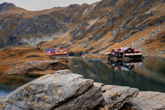 Scenic autumn landscape at Balea Lake in the Fagaras Mountains, Romania, featuring textured rocks in the foreground and a mountain cabin reflected in the calm water.