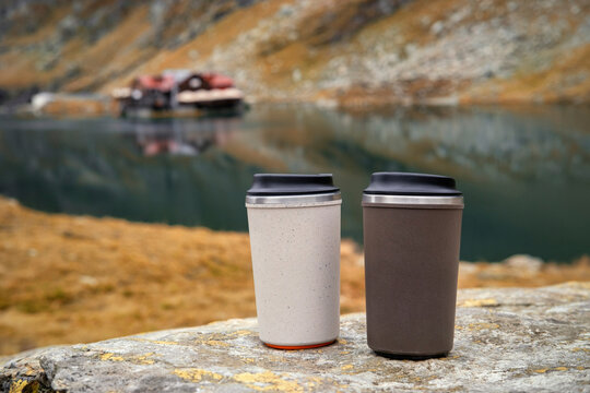 Two eco-friendly reusable travel mugs resting on a rock with the scenic autumn landscape of Balea Lake and the Fagaras Mountains, Romania, in the background.