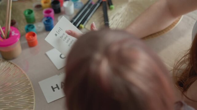 Shows adult and child engaged in phonics learning at table. Adult holds card with letter transcription while pointing to it surrounded by art supplies like paint containers