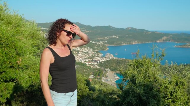 Woman suffering from heat exhaustion. Middle-aged woman in sunglasses fanning herself and feeling unwell from the intense summer heat while standing on a hill overlooking a beautiful mediterranean bay