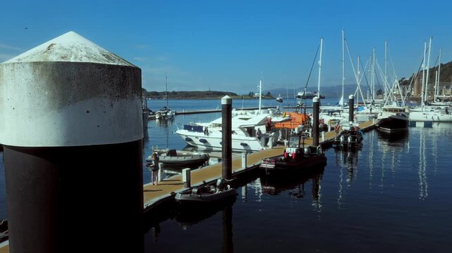 A large pier post anchors the foreground beside marina pontoons, moored yachts and rows of masts in Oban, Scotland.